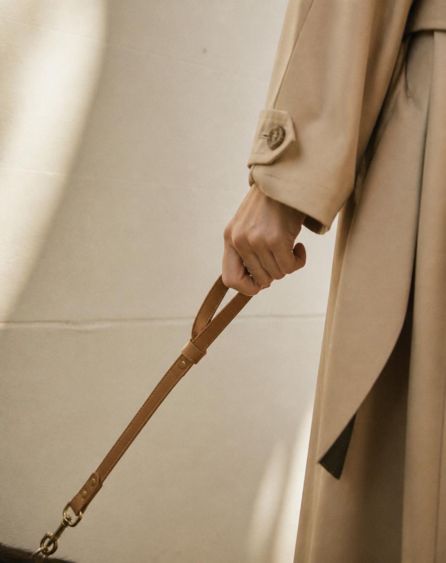 A slim tan leather CHILLI lead held in the hand of a trench coat